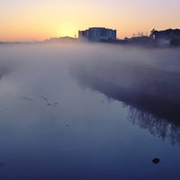 朝霧立ち込める河川敷の朝焼け、対岸の建物シルエットの写真