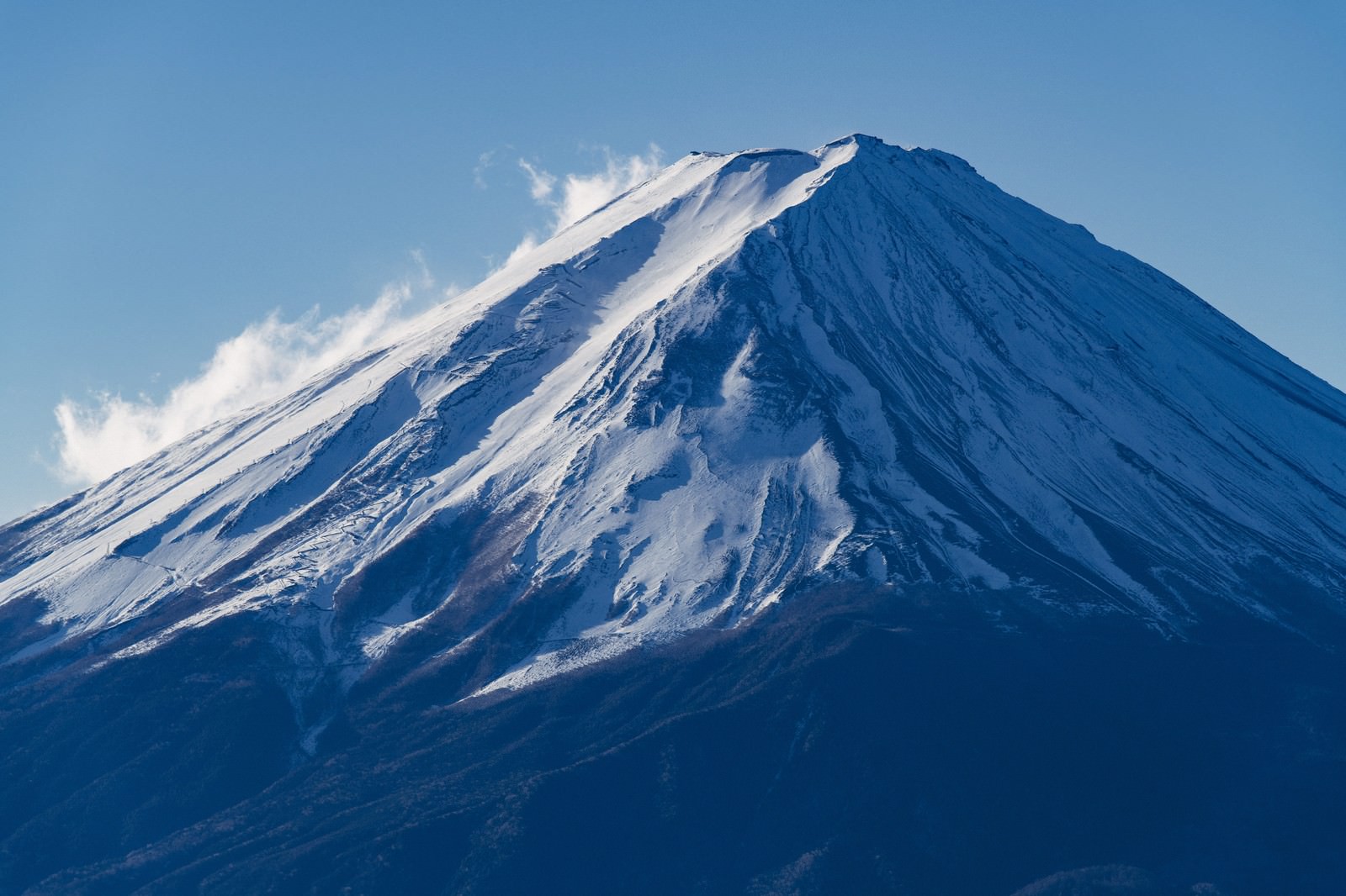 青空の下で雪化粧した富士山の山頂から白い雪煙が舞い上がる