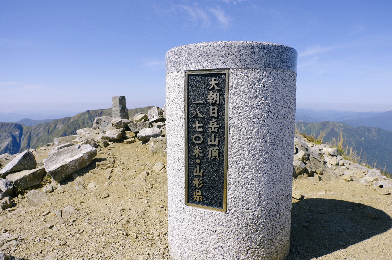 青空の下、大朝日岳山頂の岩場に立つ山頂碑と周囲の登山者たち