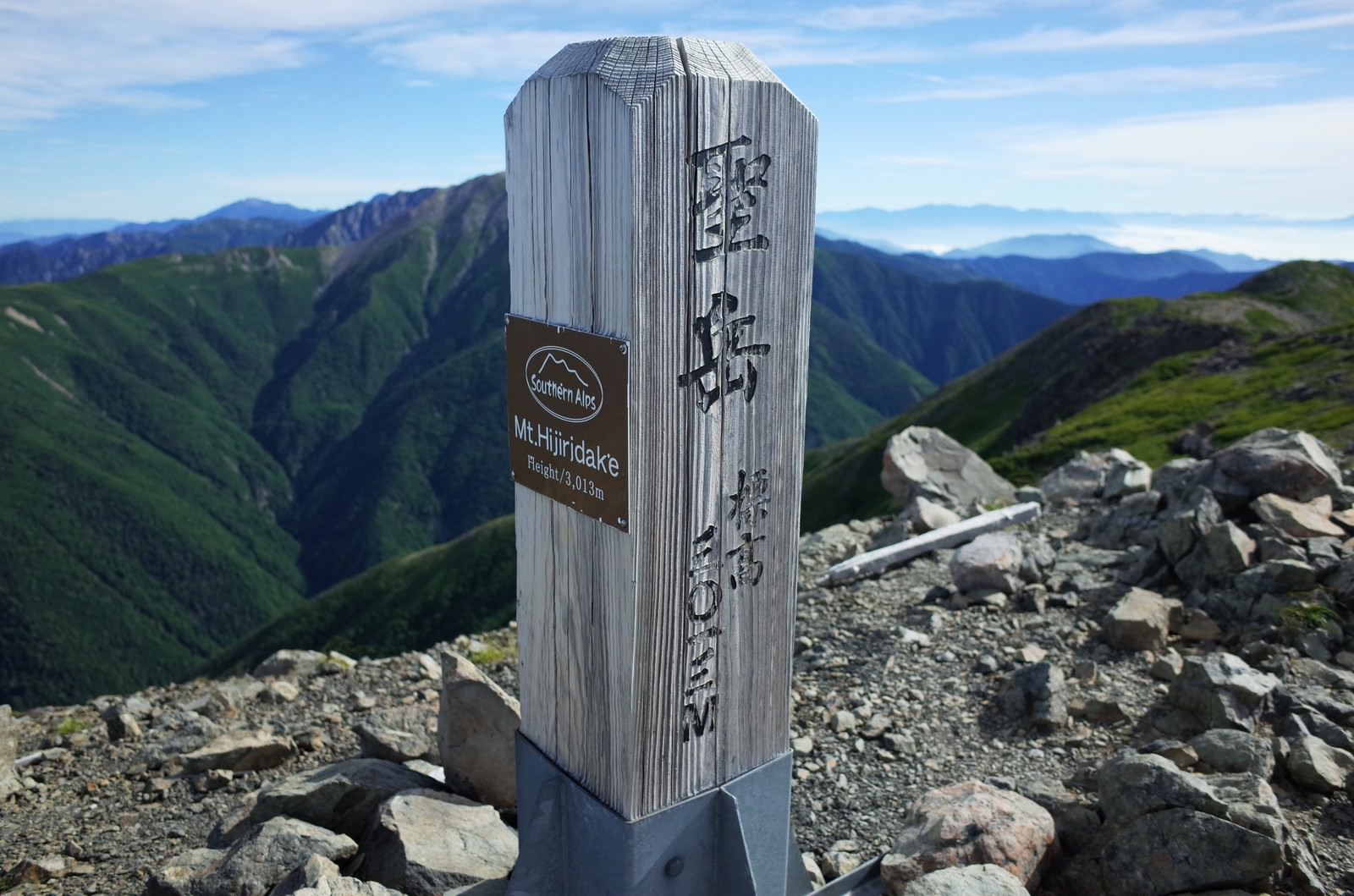 青空の下、聖岳山頂に立つ白い木製の山頂碑と背景の山々