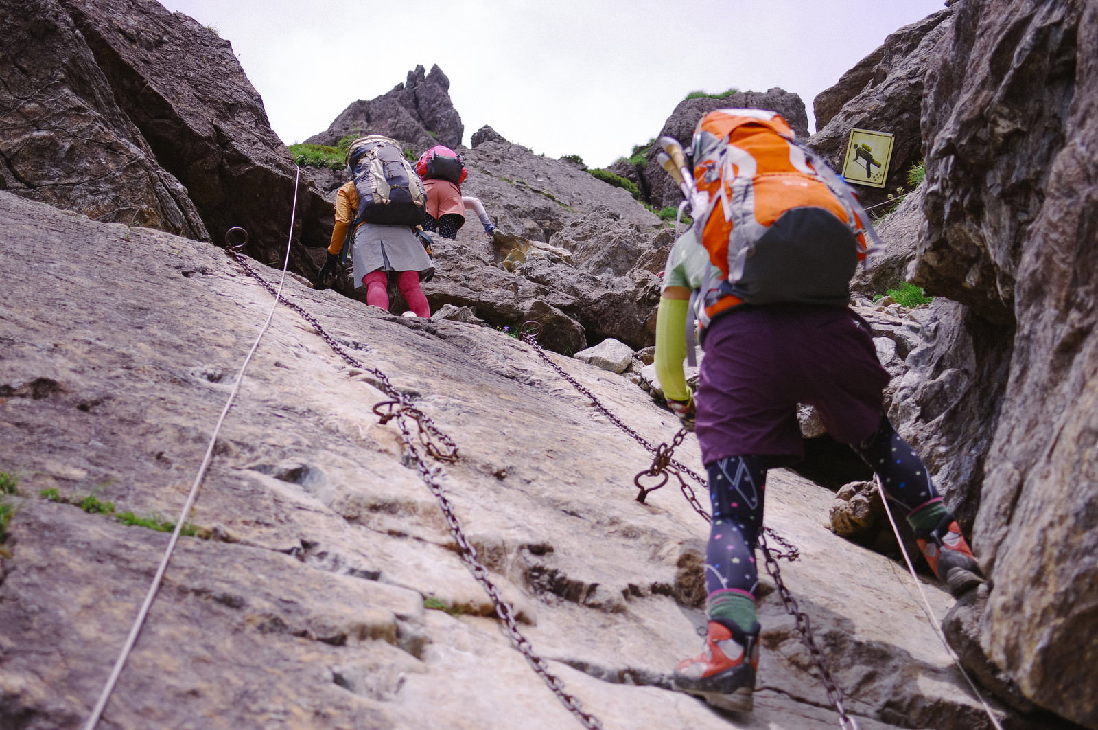 険しい岩肌の鎖場を登るオレンジ色のヘルメットを着用した登山者たち