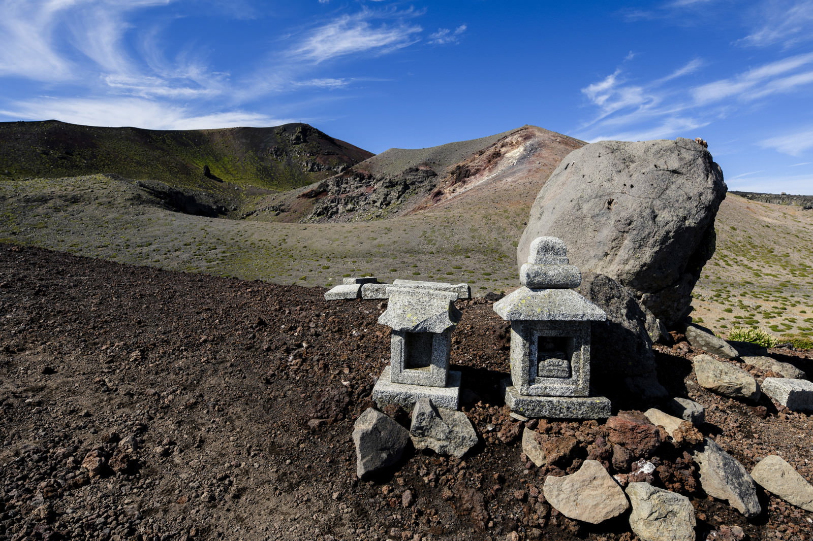 岩手山の荒涼とした山頂に石で組まれた小さな社が建ち、青空と雲が背景に広がる