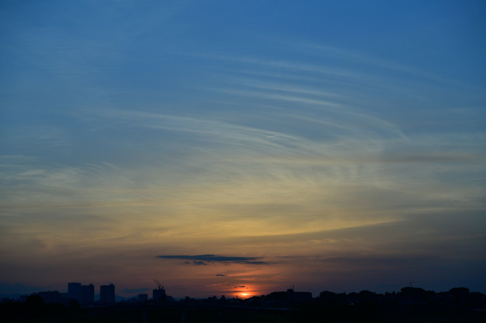 オレンジ色の夕焼け空に細い飛行機雲が一筋伸び、地平線に太陽が沈む風景