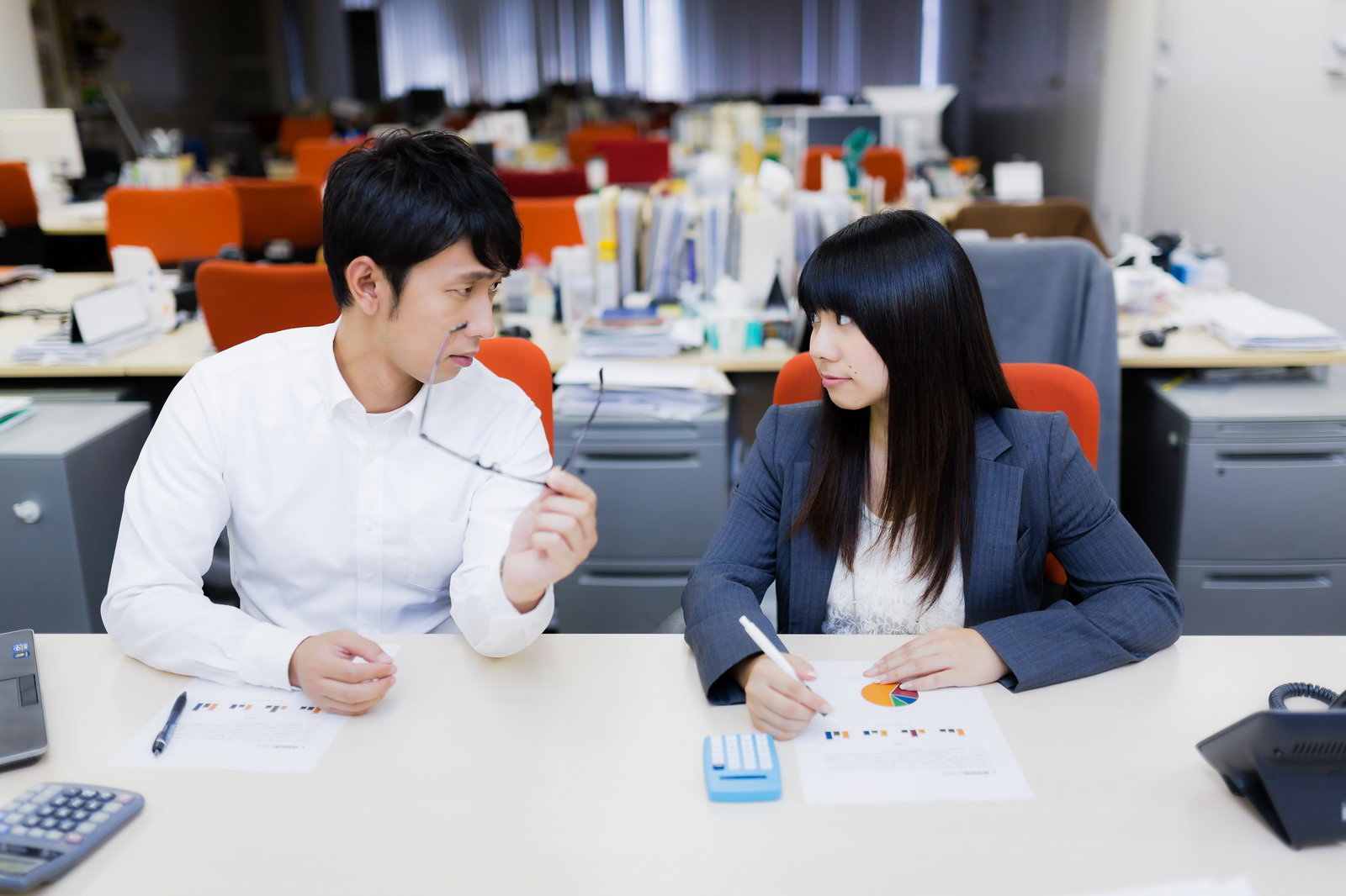 白いシャツの男性がメガネを手に笑う隣で、濃紺ジャケットの女性が書類を持ち冷めた表情で見つめている［モデル：大川竜弥 Lala］
