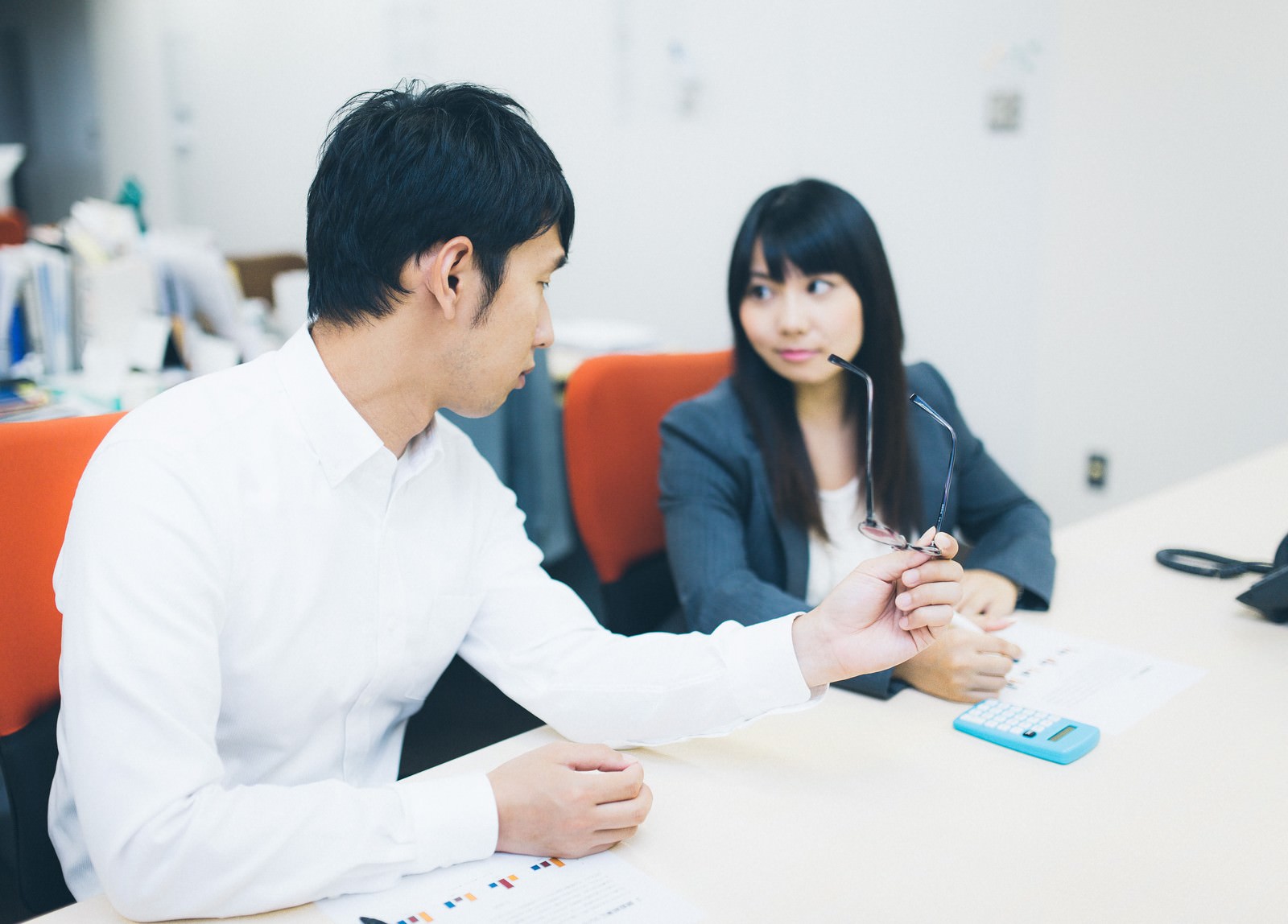 白シャツの男性が書類を持ちながら、眼鏡を手にした濃紺ジャケットの女性と机で向き合っている［モデル：大川竜弥 Lala］