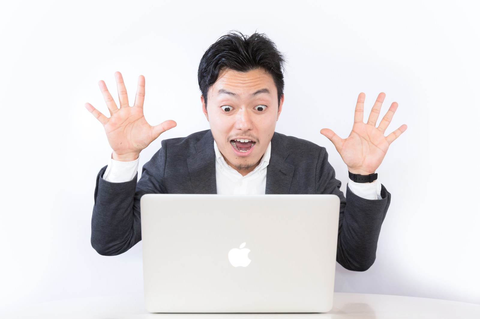 スーツの男性がノートパソコンの前で両手を上げ、驚いた表情を浮かべている［モデル：藤沢篤］