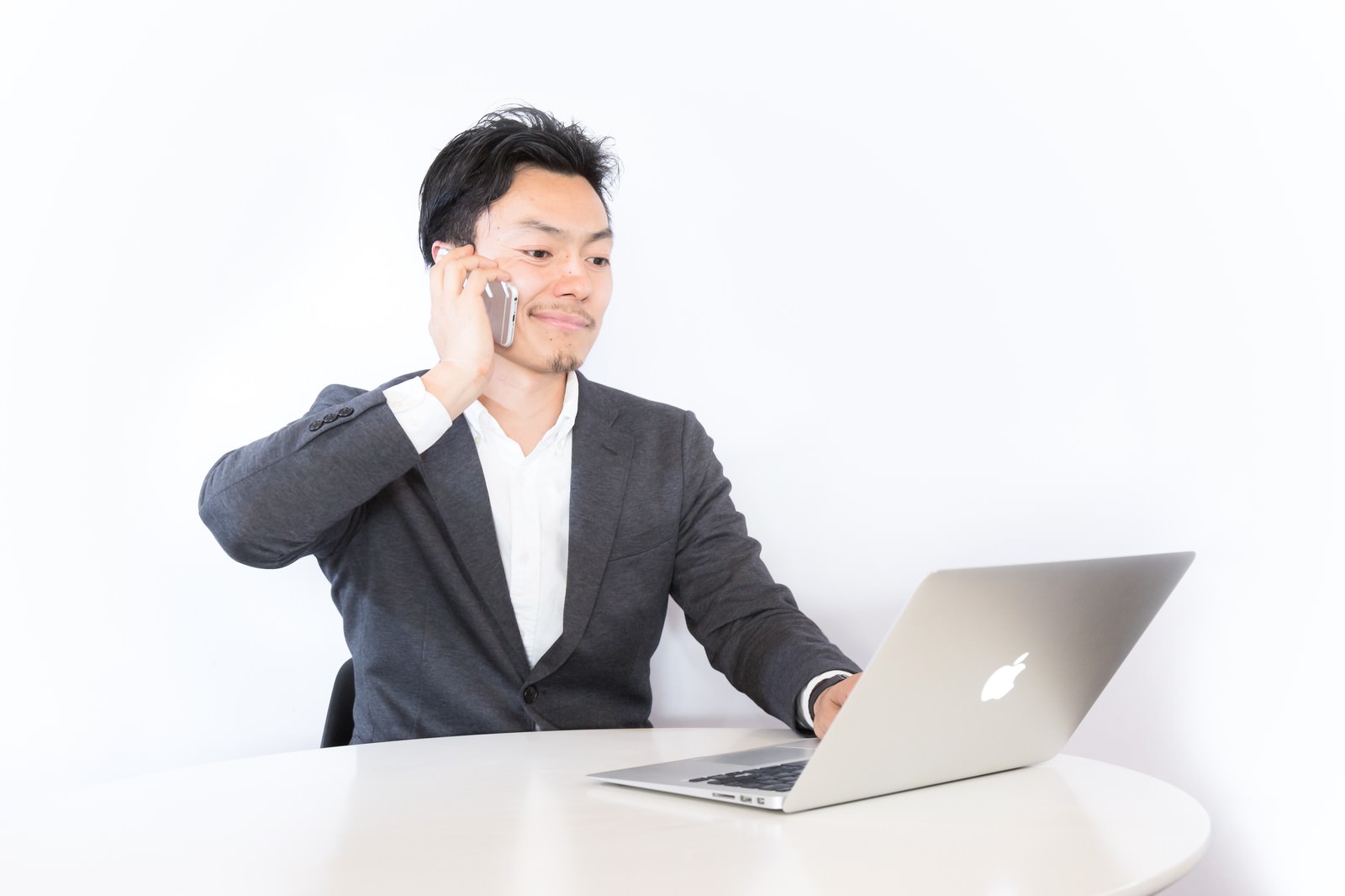 グレースーツの男性がノートパソコンを操作しながらスマートフォンで通話している