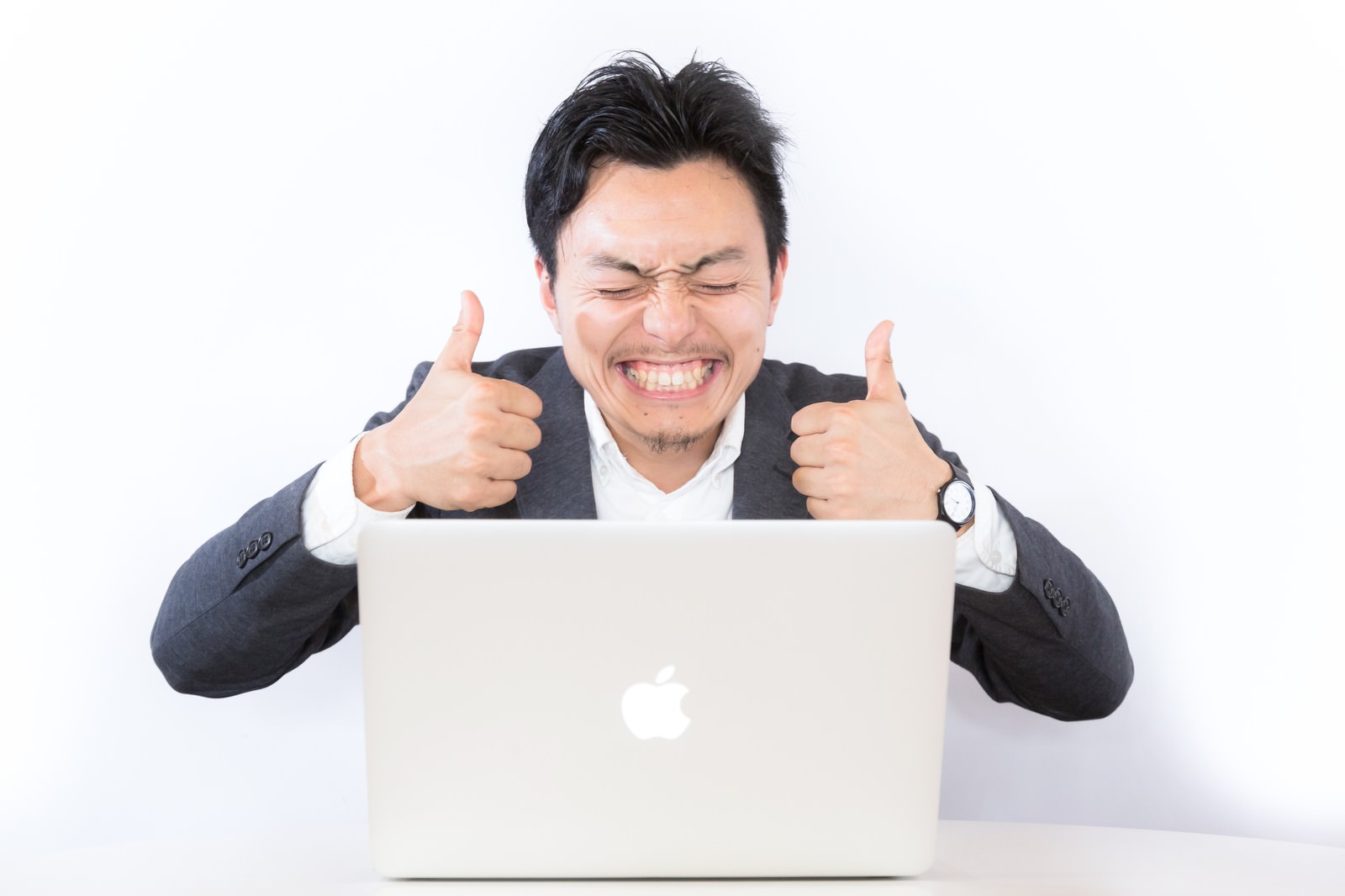 ノートパソコン前で両手サムズアップするスーツの男性［モデル：藤沢篤］