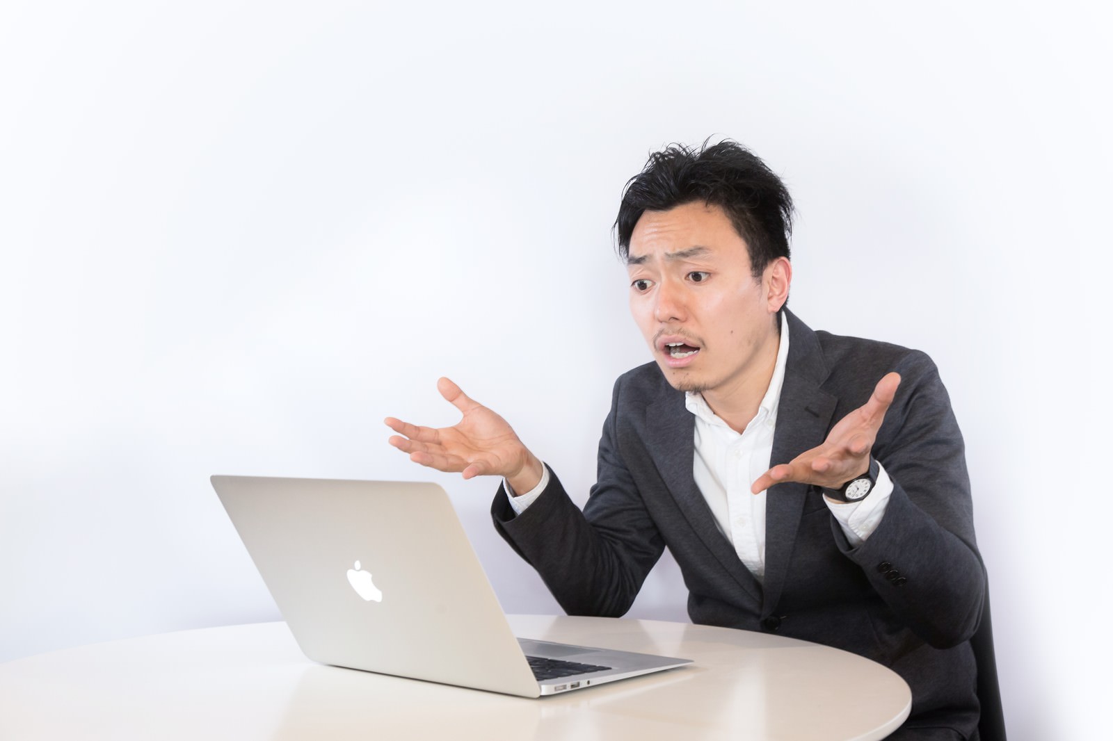 ノートパソコンの前で両手を広げて呆れるスーツ姿の男性［モデル：藤沢篤］