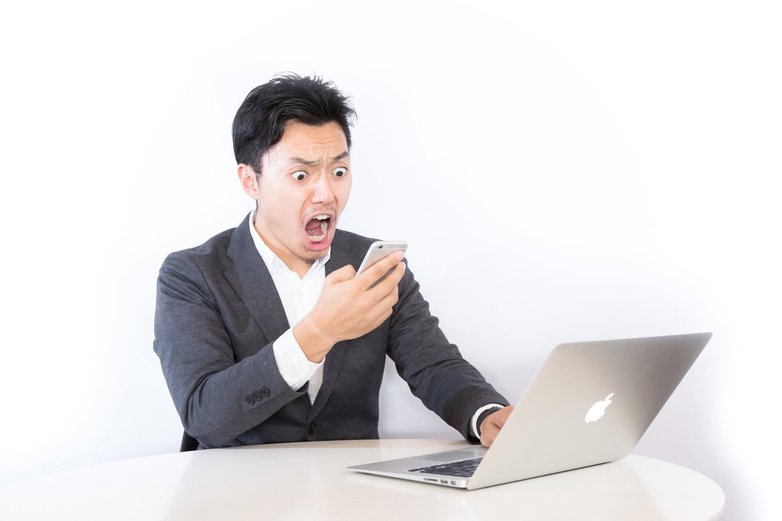 デスクでスマートフォンを見て驚くスーツ姿の男性［モデル：藤沢篤］