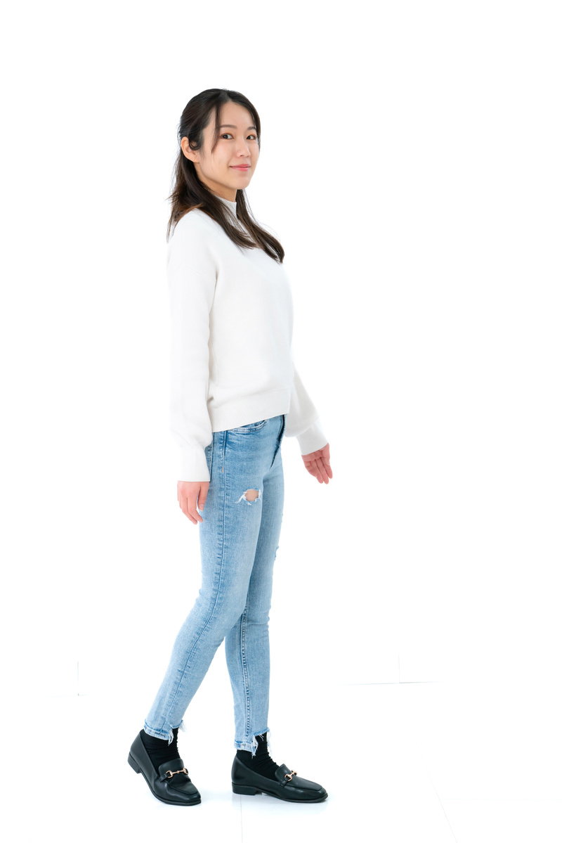 白いトップスとジーンズを着て白い背景で立つ女性［モデル：SAKI］