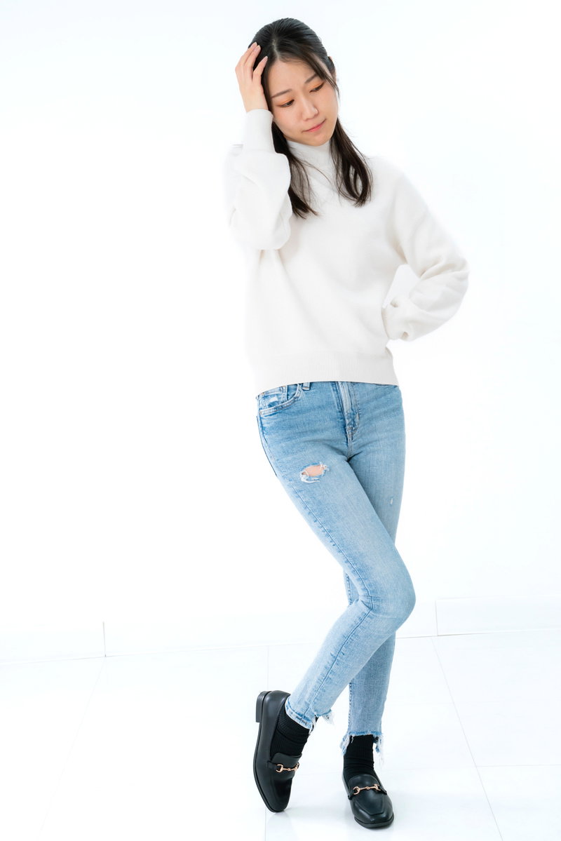 白いニットとジーンズの女性が片手を腰に当て困惑した表情で立つ［モデル：SAKI］
