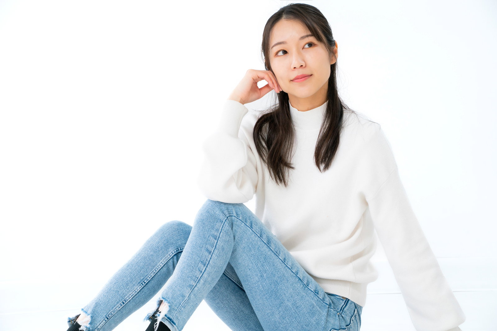 膝を立てて座り遠くを見つめる白いトップス姿の女性［モデル：SAKI］