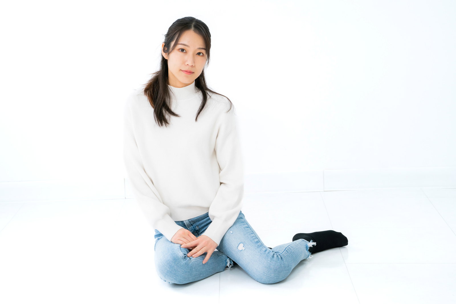 白いニットとデニムパンツを着た女性が白背景で横座りのポーズ［モデル：SAKI］