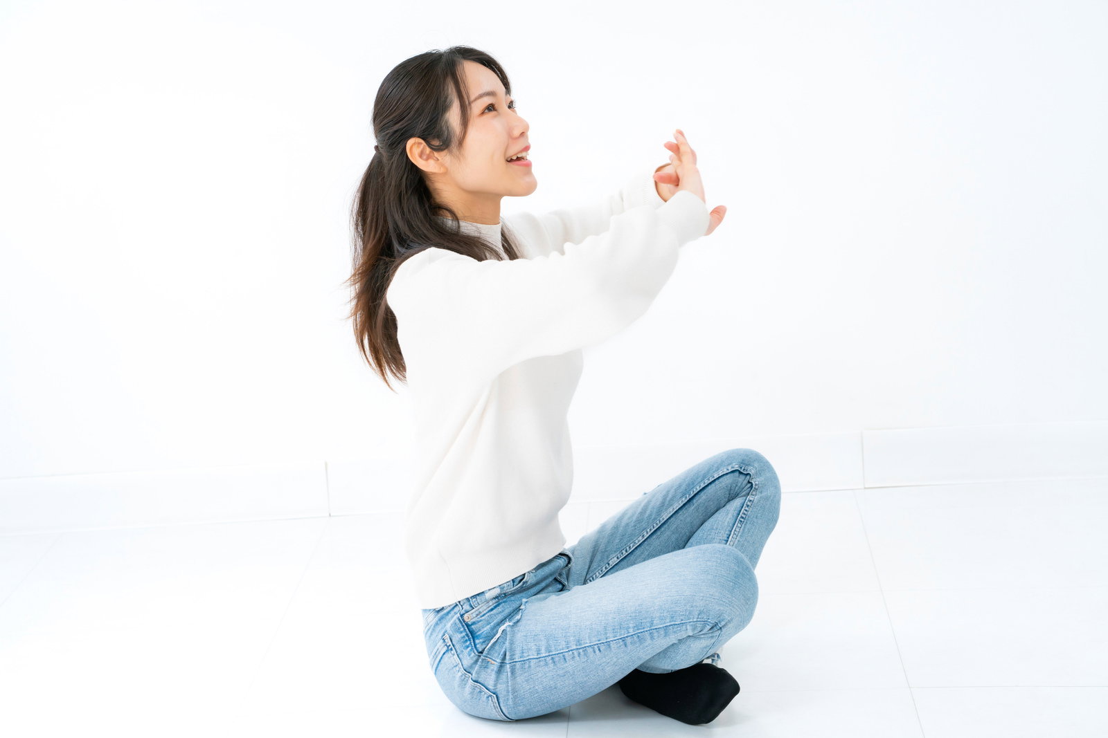 白背景であぐら座りをしながら上半身をストレッチする女性［モデル：SAKI］