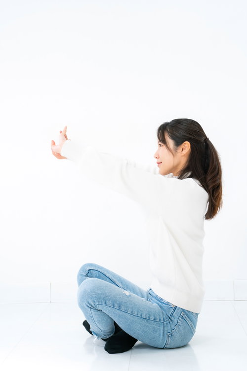 白背景であぐら座りで片腕を前方に伸ばしてストレッチする女性