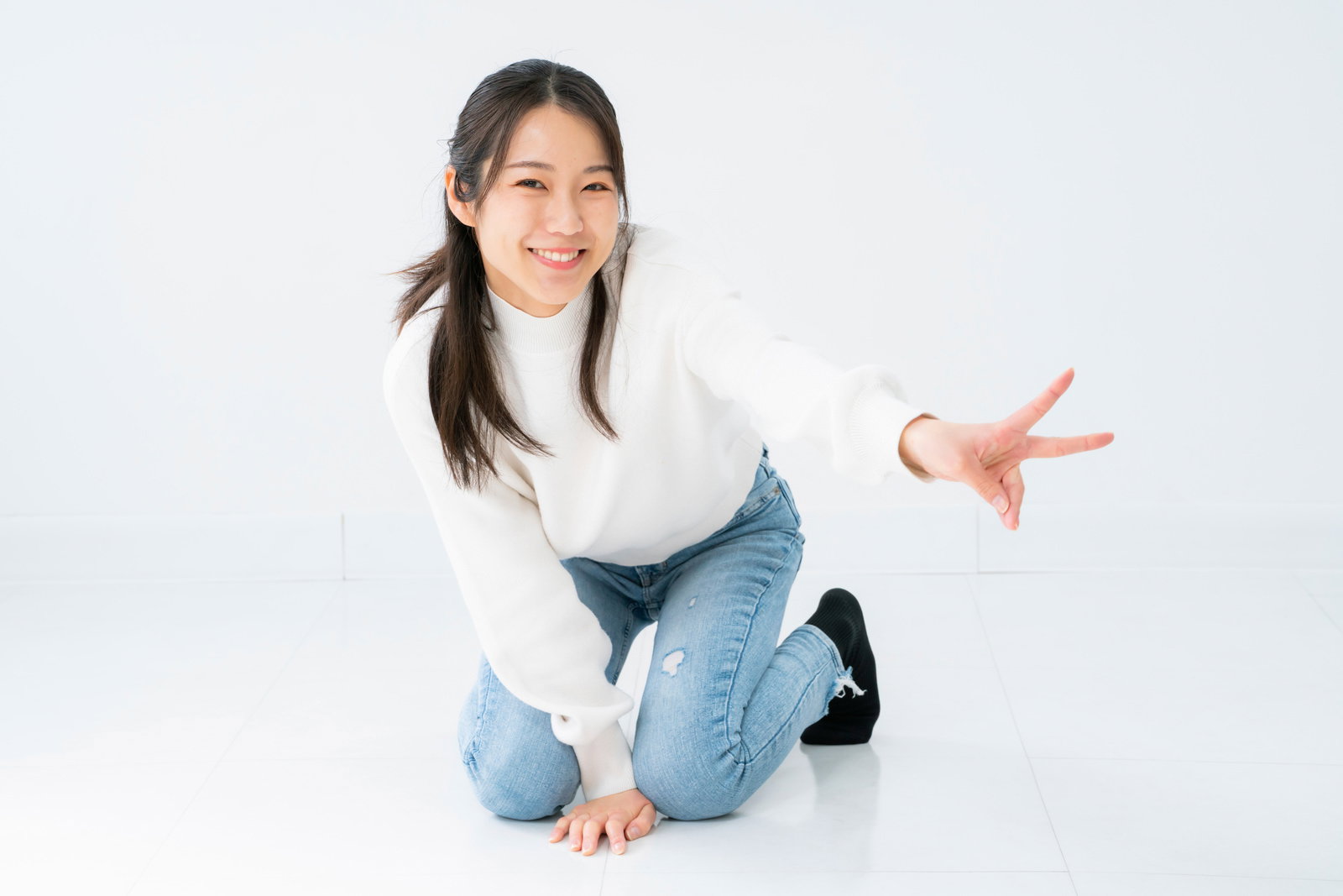 白背景で膝立ちしてピースする笑顔の女性［モデル：SAKI］