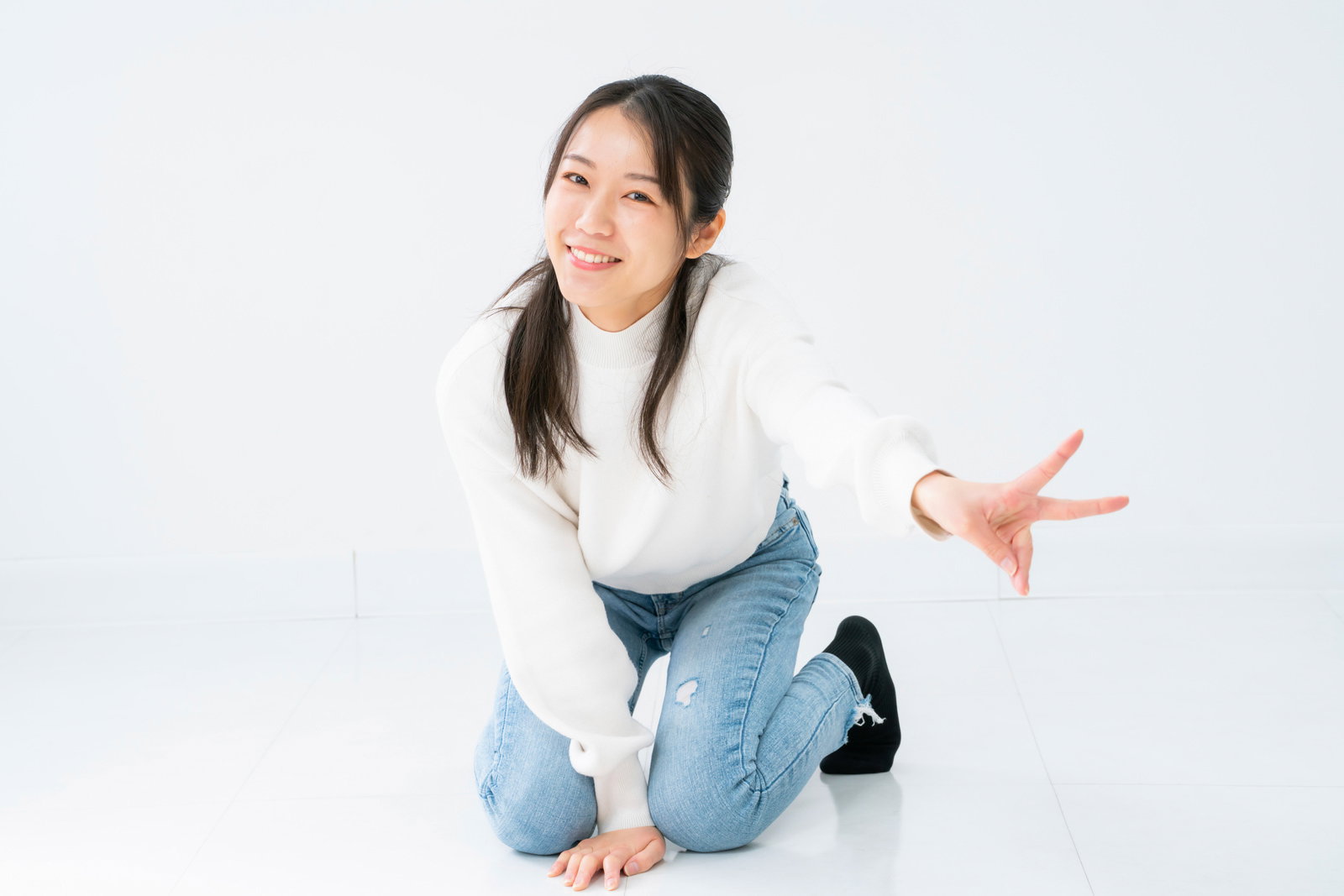白背景で膝立ちしながらピースサインを送る女性［モデル：SAKI］