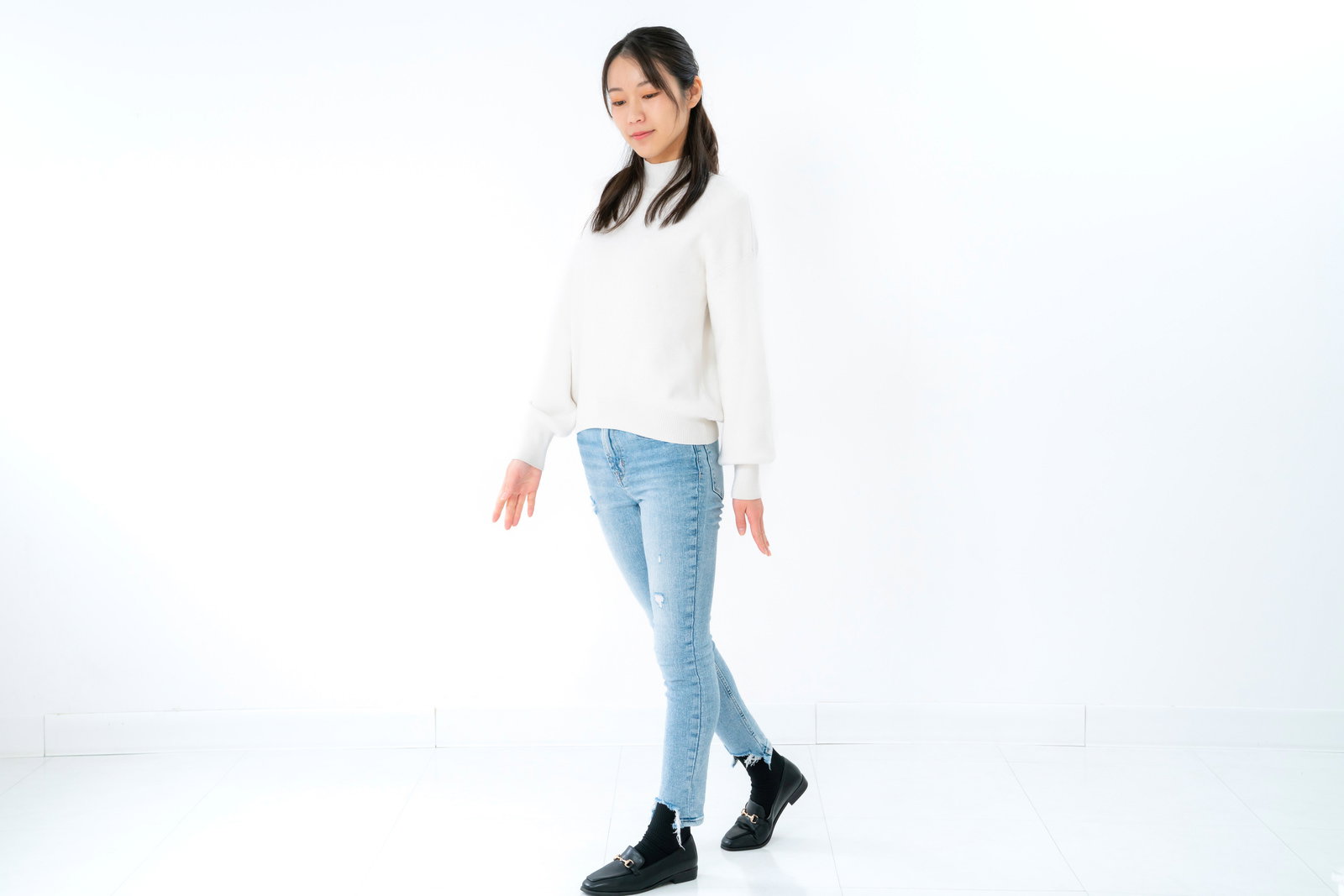 白いトップスと淡いデニム、黒いブーツを着た女性が伏し目がちに立ち止まる全身ポーズ［モデル：SAKI］