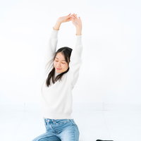 白背景で横座りして両腕を上げて伸びをする女性の写真