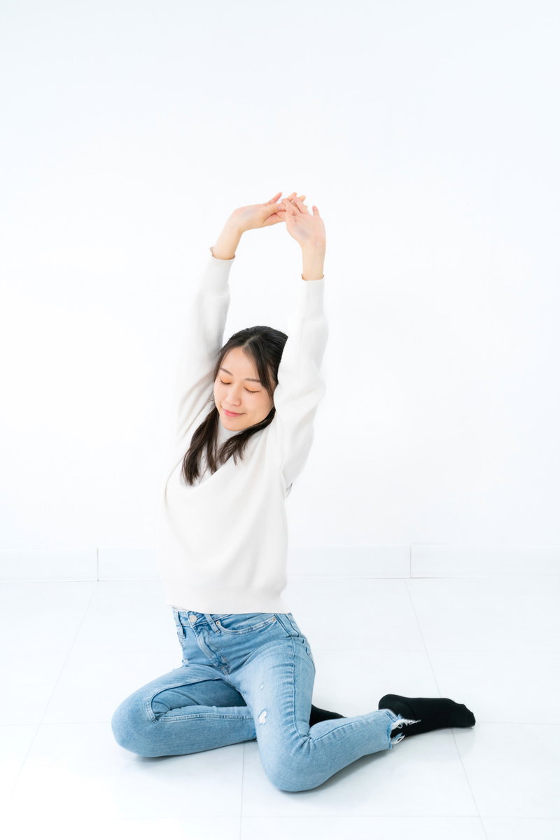 白背景で横座りして両腕を上げて伸びをする女性［モデル：SAKI］