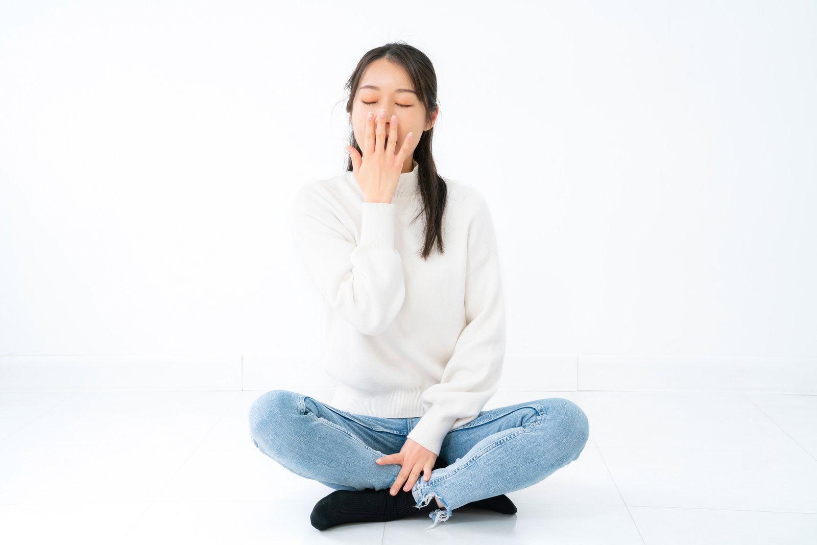 白いニットを着た女性が床に座り手を口元に当ててあくびをしている［モデル：SAKI］