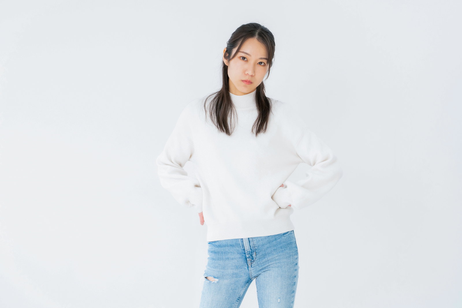 腰に手を当てて不機嫌な表情で睨む白いトレーナー姿の女性［モデル：SAKI］