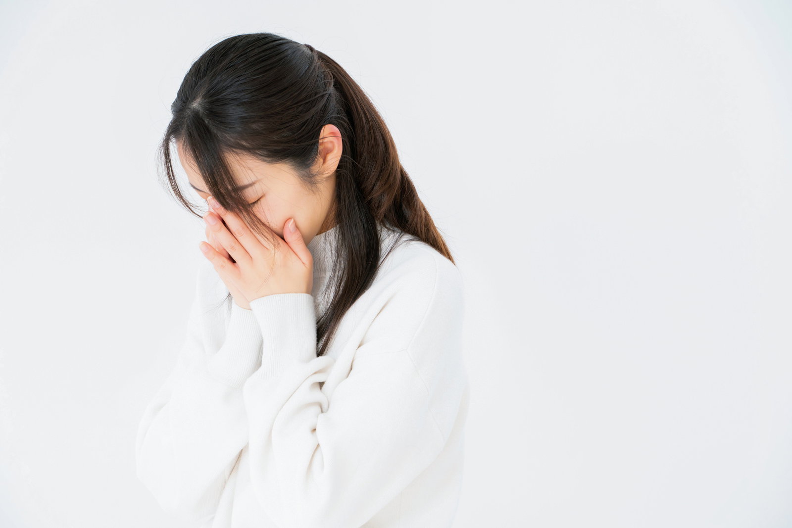 白い背景の前で、両手で顔を覆いながら泣く女性の姿［モデル：SAKI］