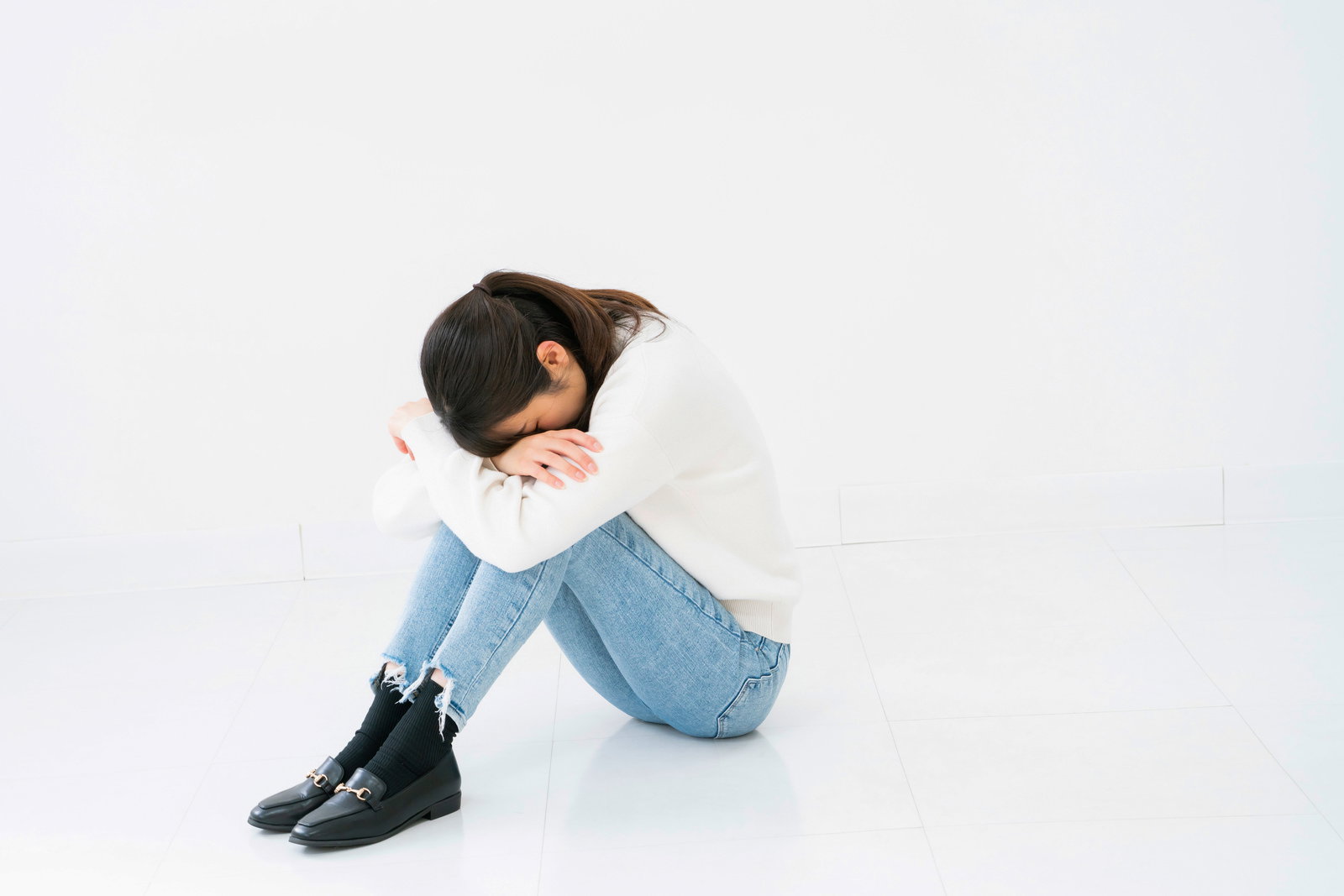 白い背景で小山座りをして項垂れている女性［モデル：SAKI］