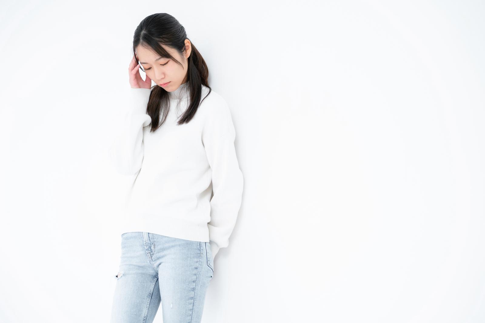 白いスウェットを着た女性が頭痛で伏し目がちに手を頭に当てている［モデル：SAKI］