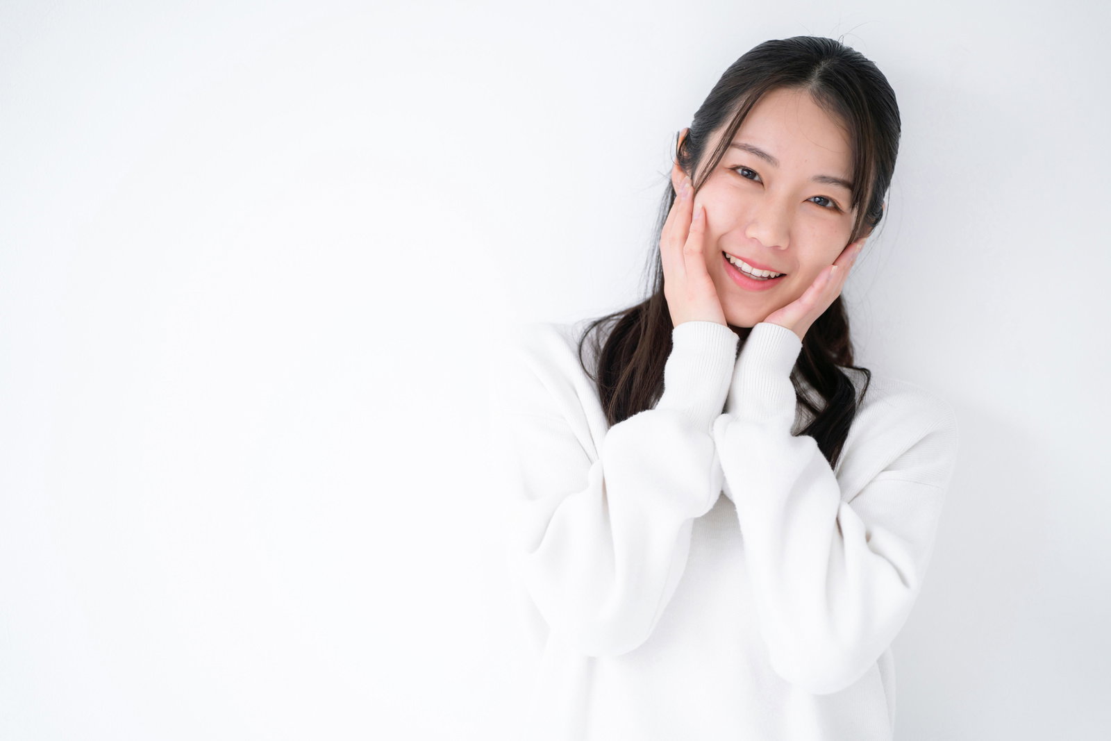 白背景で両手を頬に当てて微笑む女性のポーズ写真［モデル：SAKI］