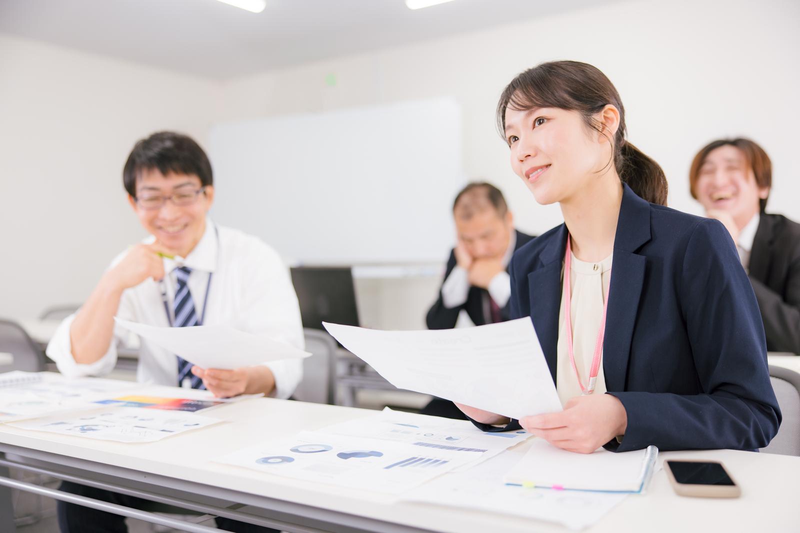 職場のミーティング室で書類を持ち確認する紺色ジャケットの女性と同僚たち［モデル：SAKI］