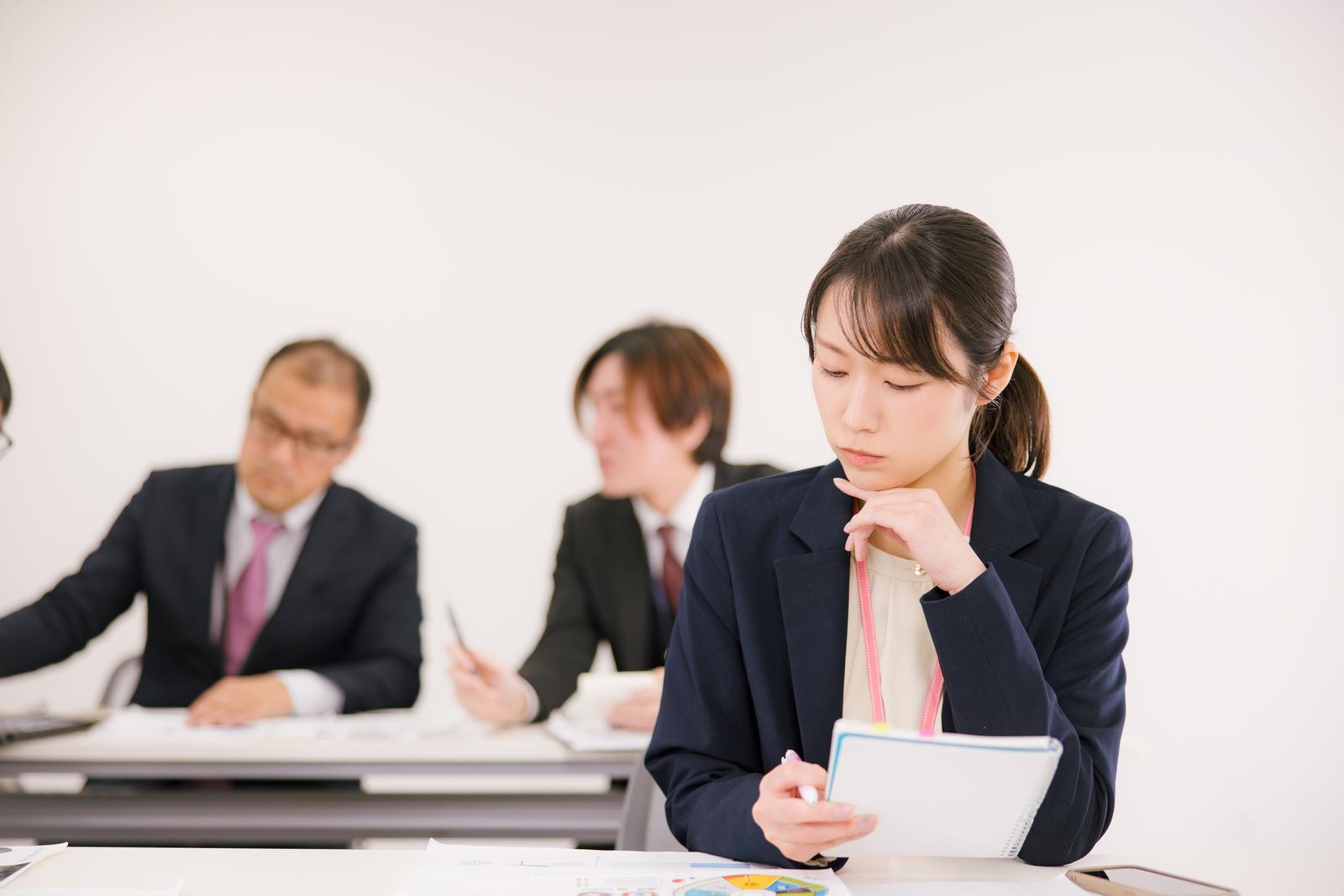 会議室で書類を持ちながら考え込む紺色スーツの女性会社員［モデル：SAKI］