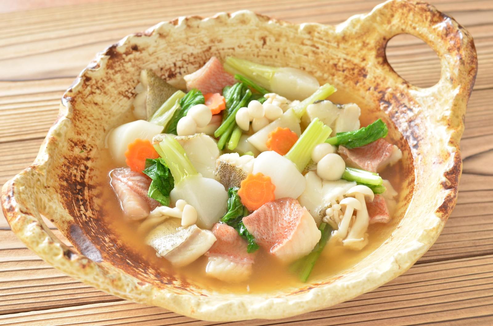 木製の器に盛られたタラと赤魚の鍋に野菜と貝類が入っている