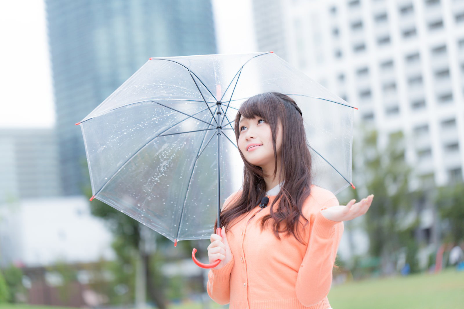 オレンジ色のシャツを着た女性が傘を持ってビル群の背景で空を見上げている［モデル：茜さや］