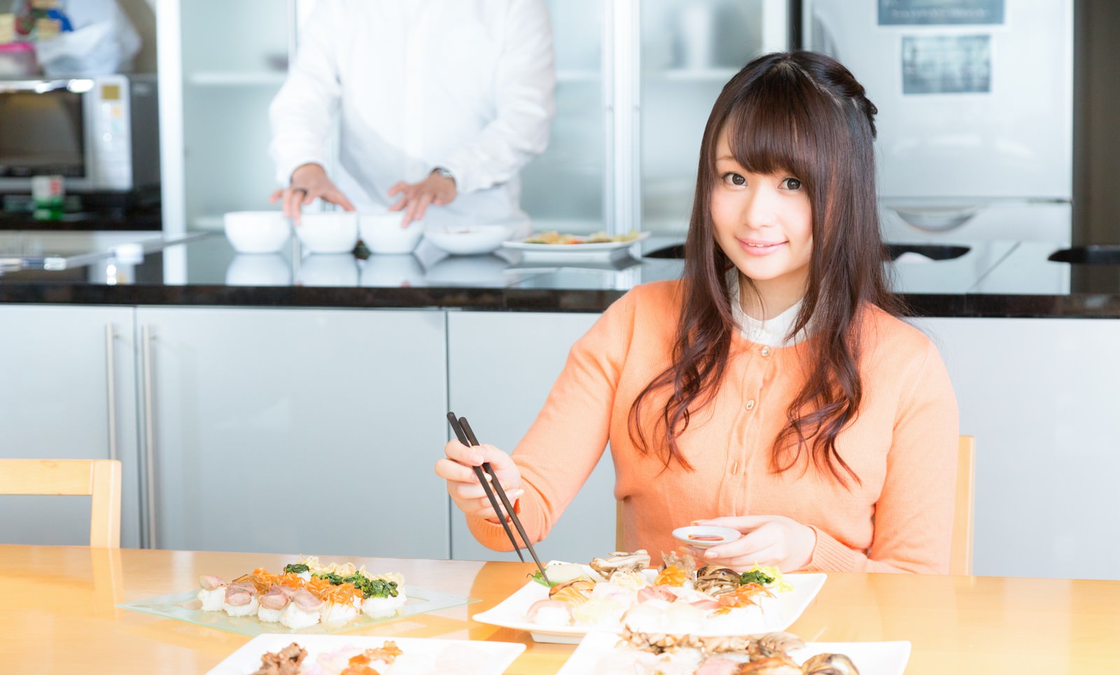 オレンジシャツの女性が食レポ中、テーブルの料理に箸を向けている［モデル：茜さや］