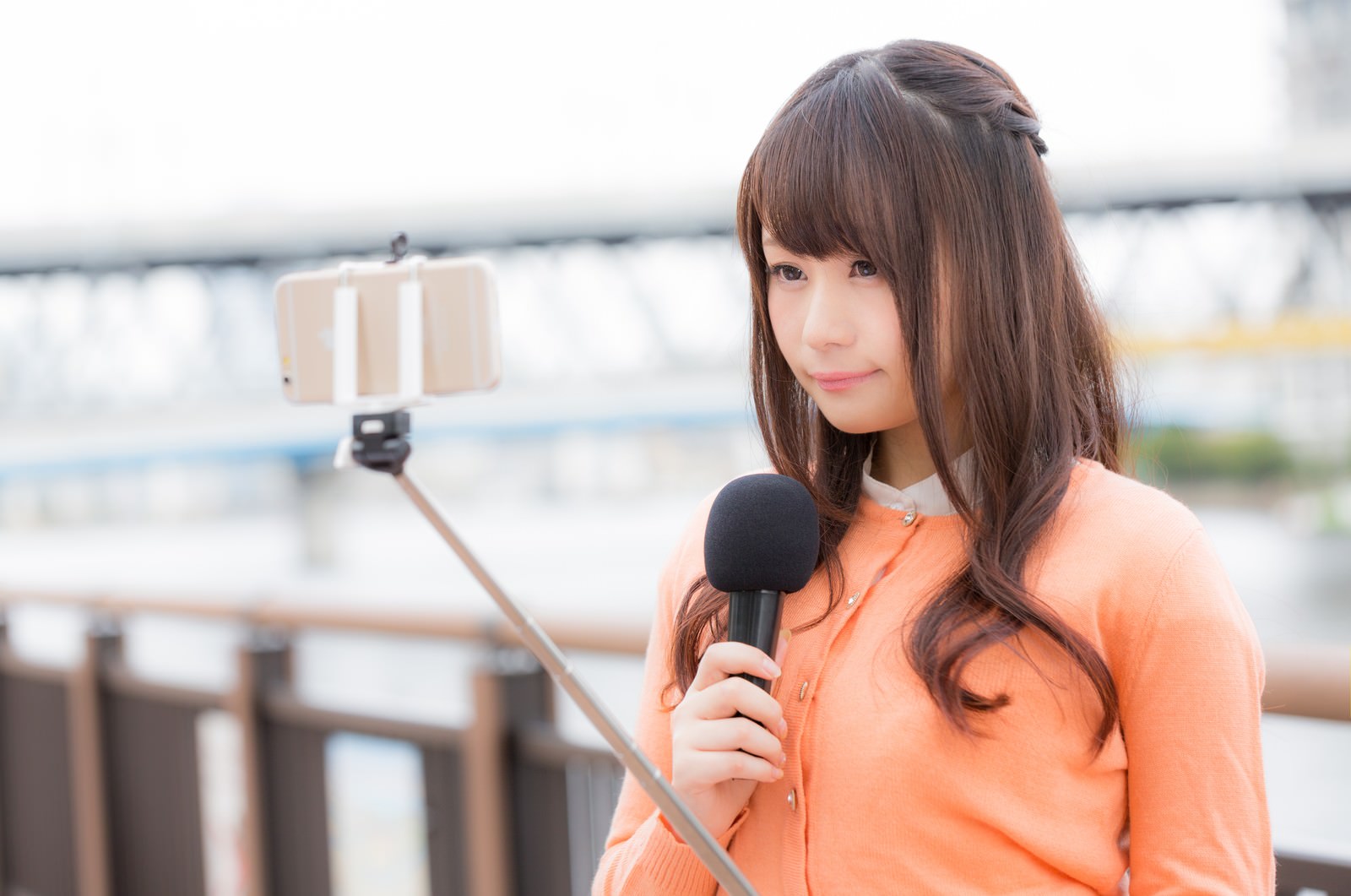オレンジシャツの女性キャスターがマイクを持ち自撮り棒でスマートフォンを固定して現場配信している［モデル：茜さや］