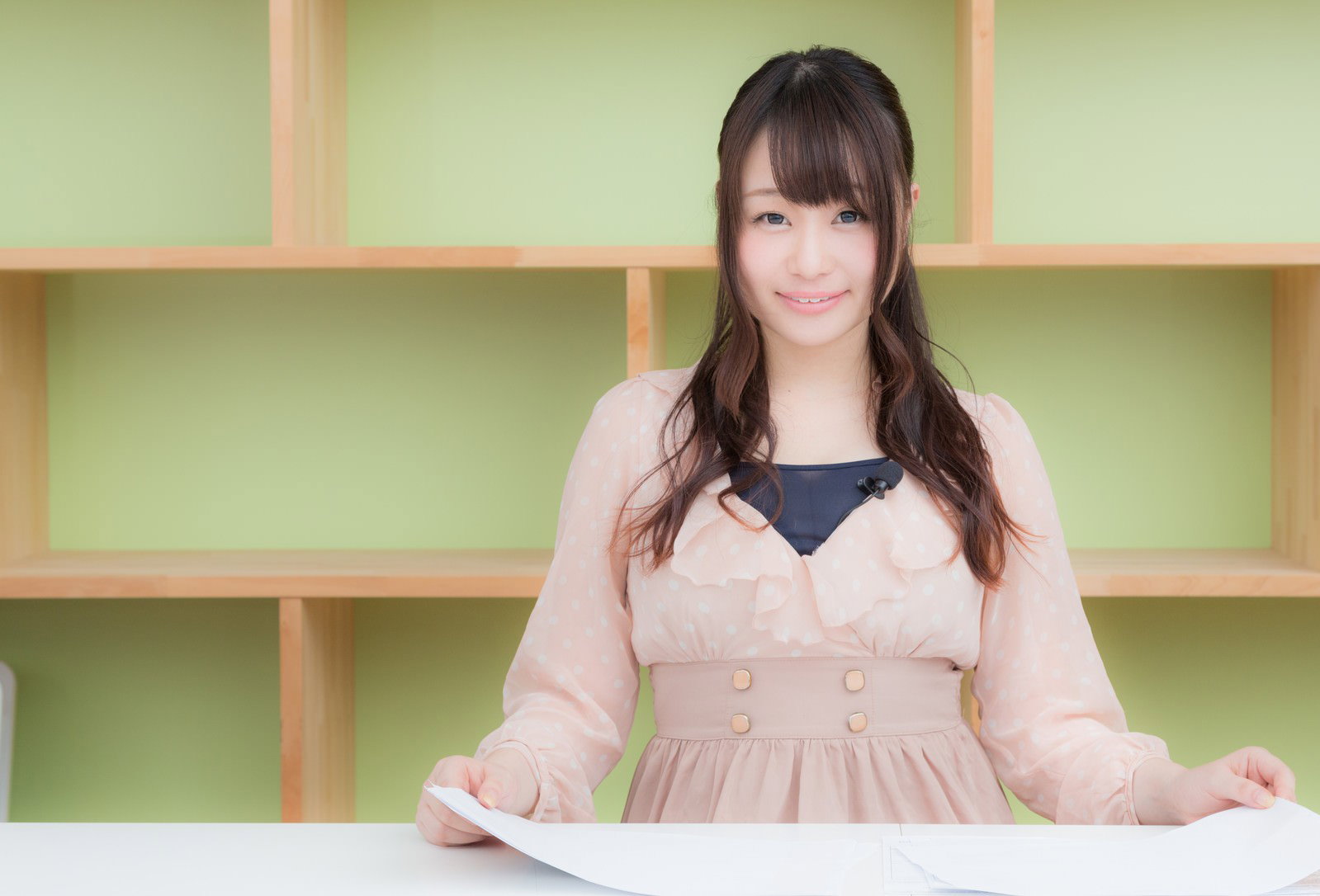 緑色の背景前でニュースを伝えるベージュのカーディガンを着た女性アナウンサー［モデル：茜さや］