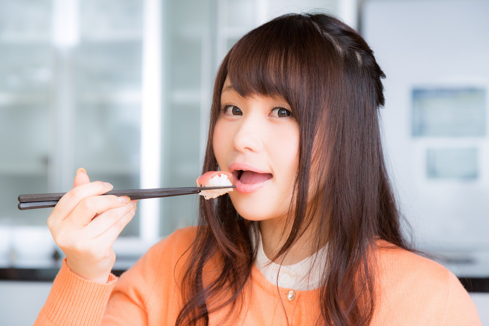 箸で握り寿司を頬張る女性がオレンジ色のトップスを着て笑顔で食べている［モデル：茜さや］