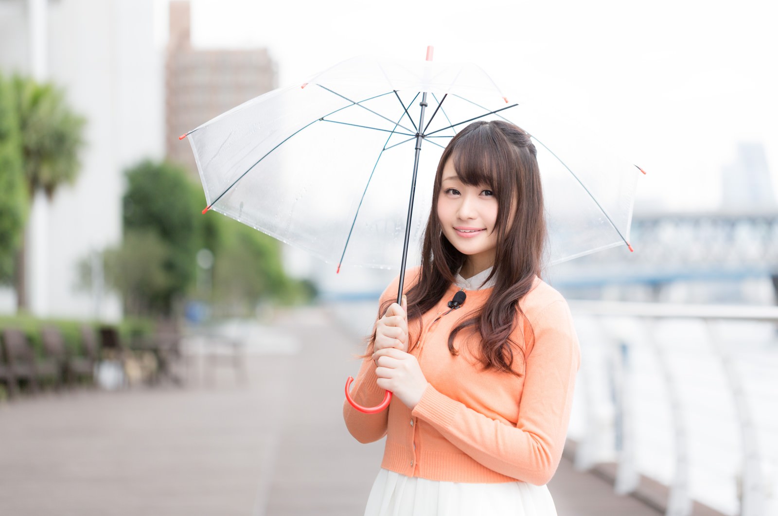 オレンジ色のカーディガン姿でグレーの傘をさし、雨の中で微笑むキャスター