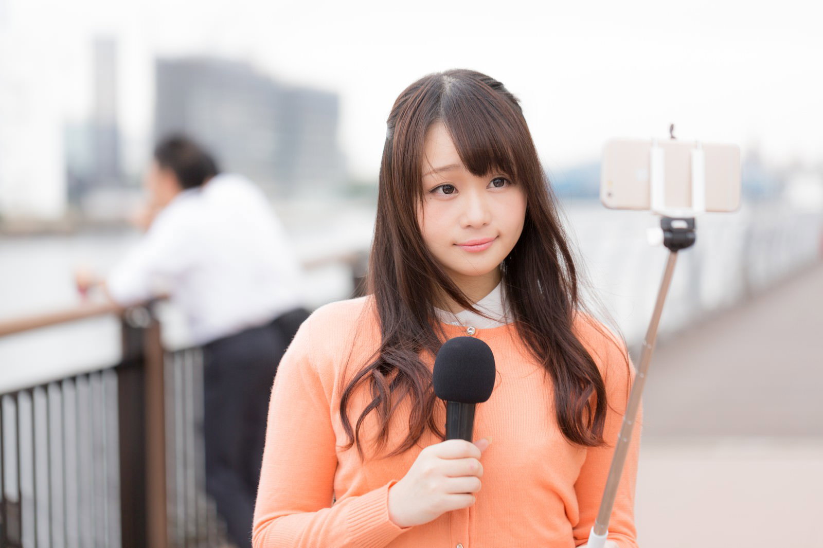 スマホと自撮り棒を持つ女性アナウンサーが屋上で現地取材を行っている［モデル：茜さや］