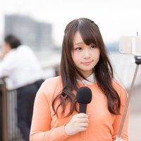 スマホと自撮り棒で現地取材を行う女性アナウンサーの写真