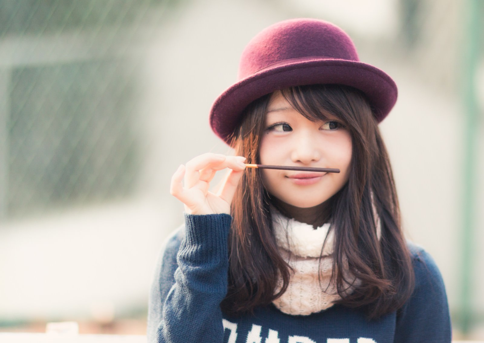 ピンク帽子をかぶった女の子がポッキーをくわえてひげに見立て、おどけた笑顔をしている［モデル：茜さや］