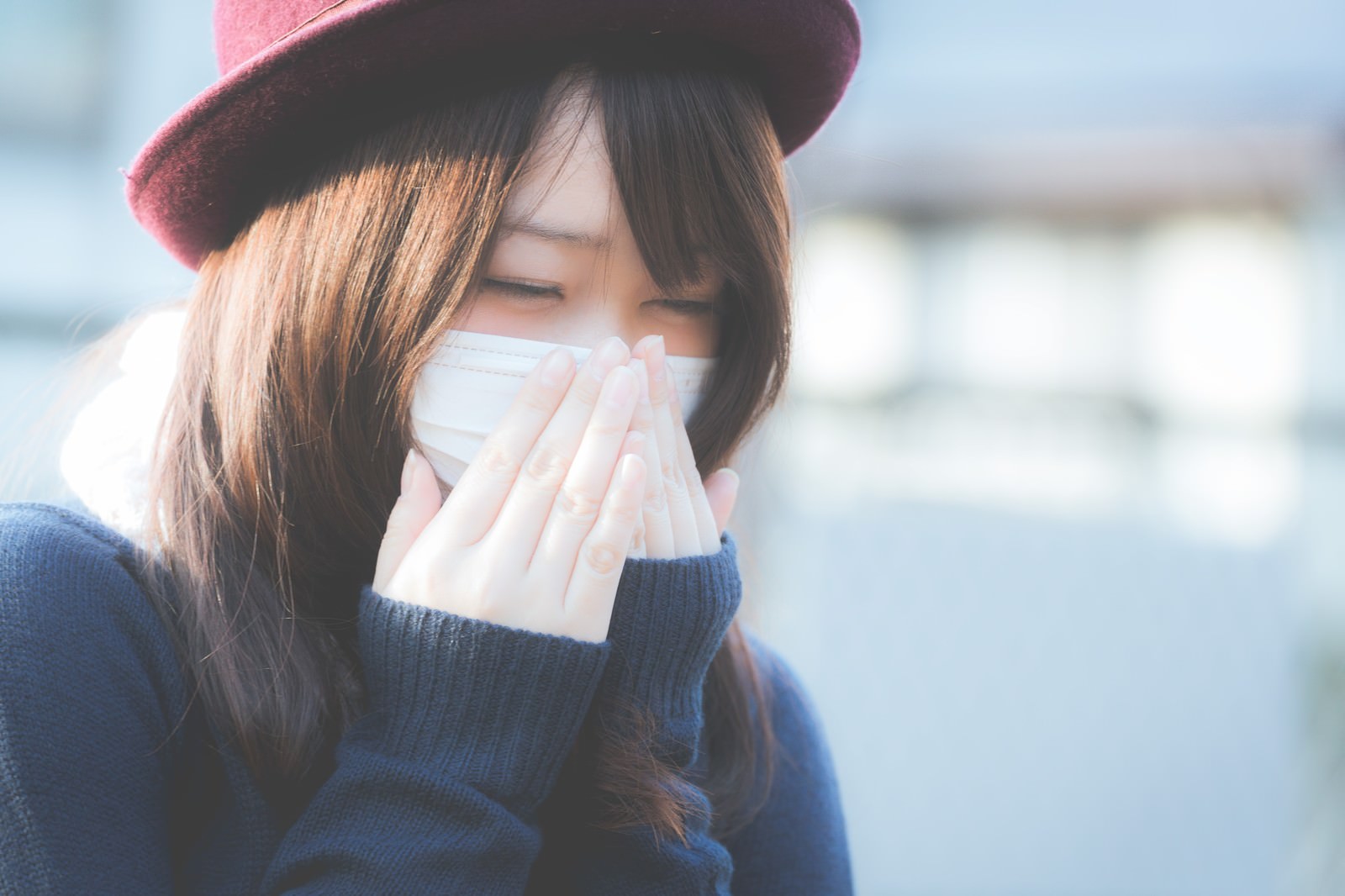 ピンクの帽子をかぶり、マスクをしてティッシュを持つ女性がくしゃみをしている［モデル：茜さや］