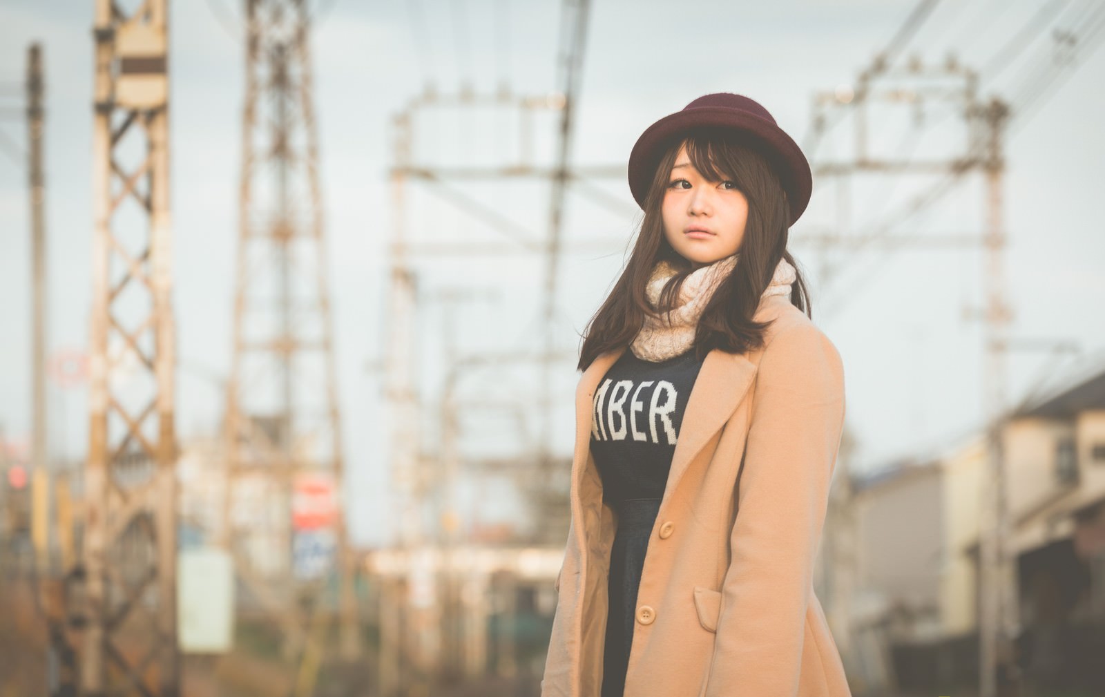 キャメルコートと帽子を着た女性が工業地帯で遠くを見つめている［モデル：茜さや］