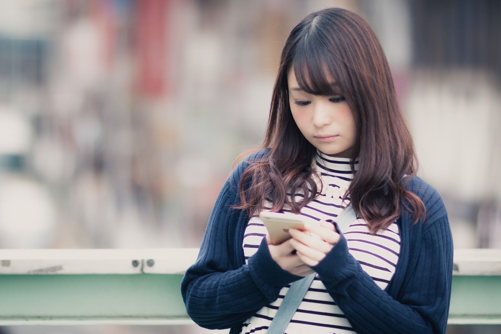 紫のストライプシャツと紺色のカーディガンを着た女性がスマートフォンで待ち合わせ場所を確認している［モデル：茜さや］