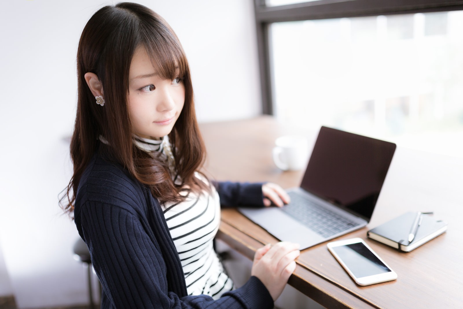 カフェで開いたパソコンとスマートフォンの前に座る女性［モデル：茜さや］