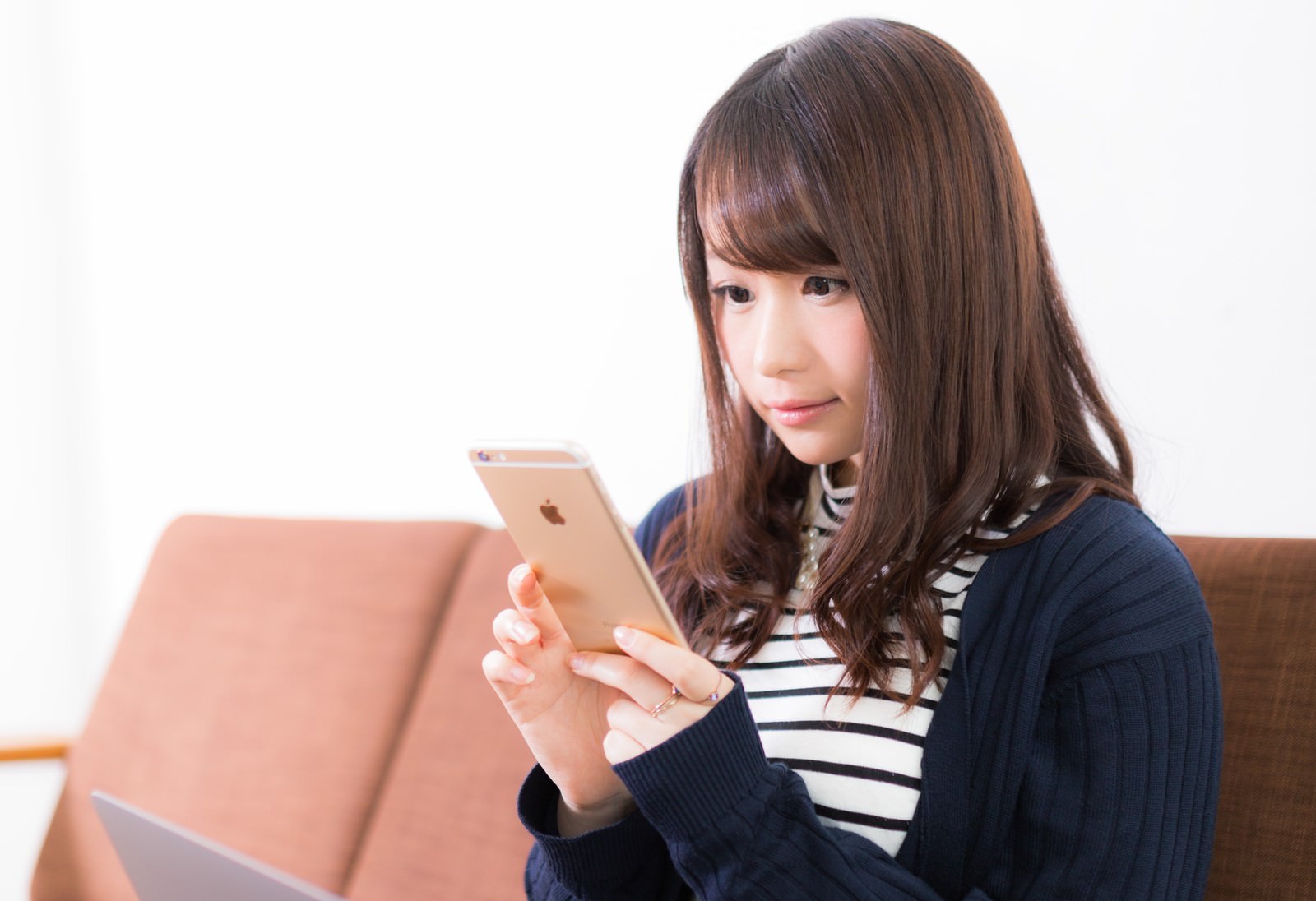 スマホで料理動画を見つめる女性がベージュのソファーに座っている［モデル：茜さや］