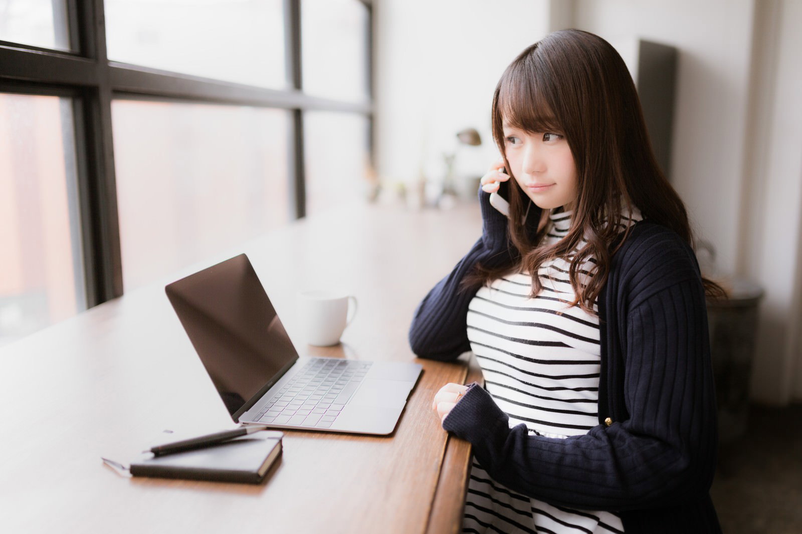 カフェで電話をするIT女性がノートパソコンの前に座っている［モデル：茜さや］