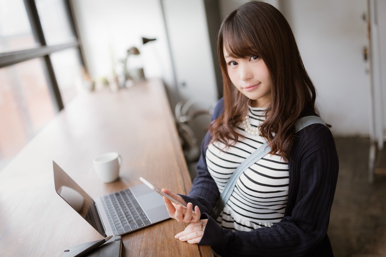 カフェのテーブルでスマートフォンを手に持ち画面を見つめるボーダー柄トップス姿の女性［モデル：茜さや］